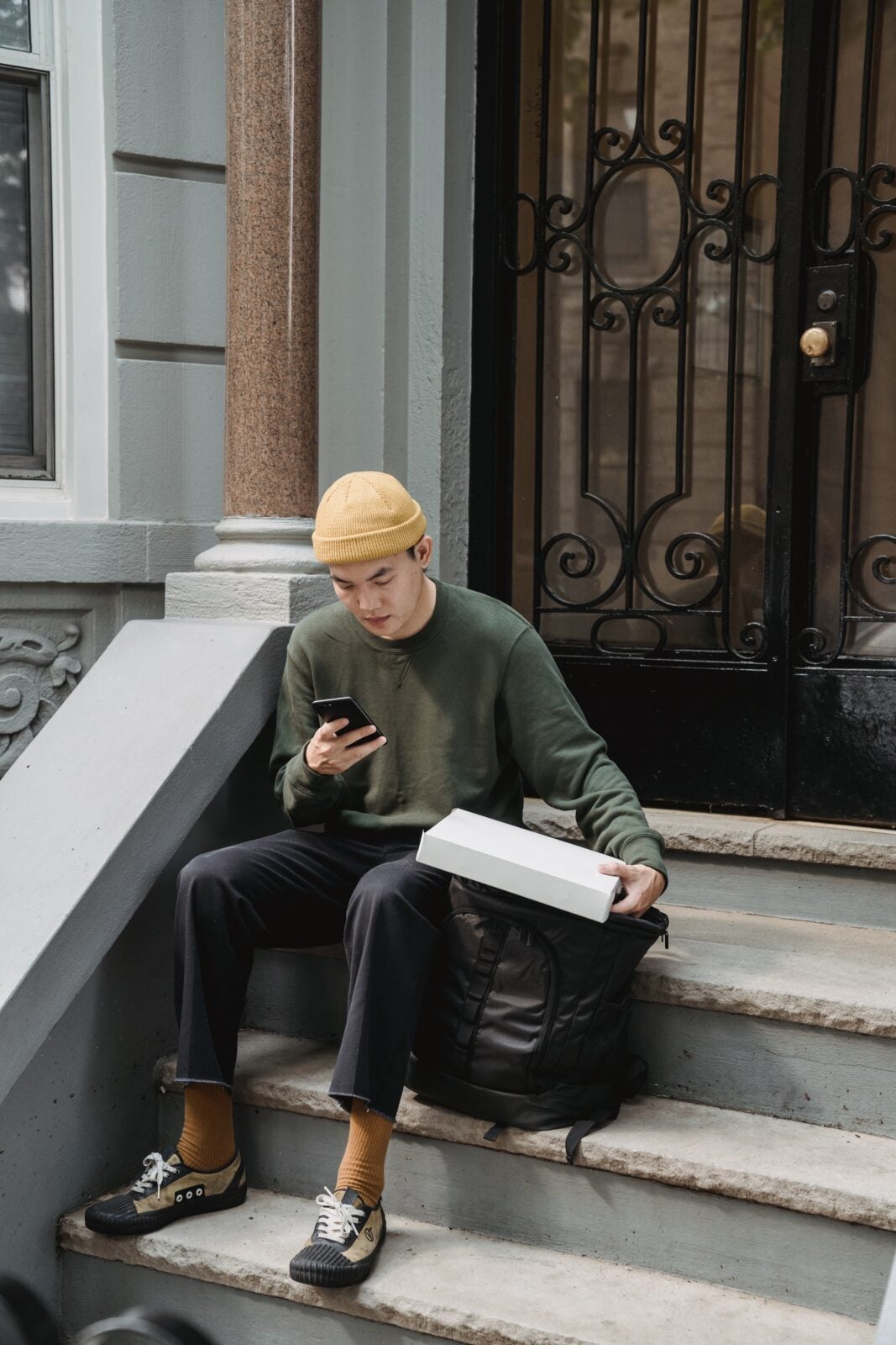 A young Asian man wearing a yellow beanie and green sweater sits at a staircase in front of a home while looking at his phone and holding a white package in his left hand.