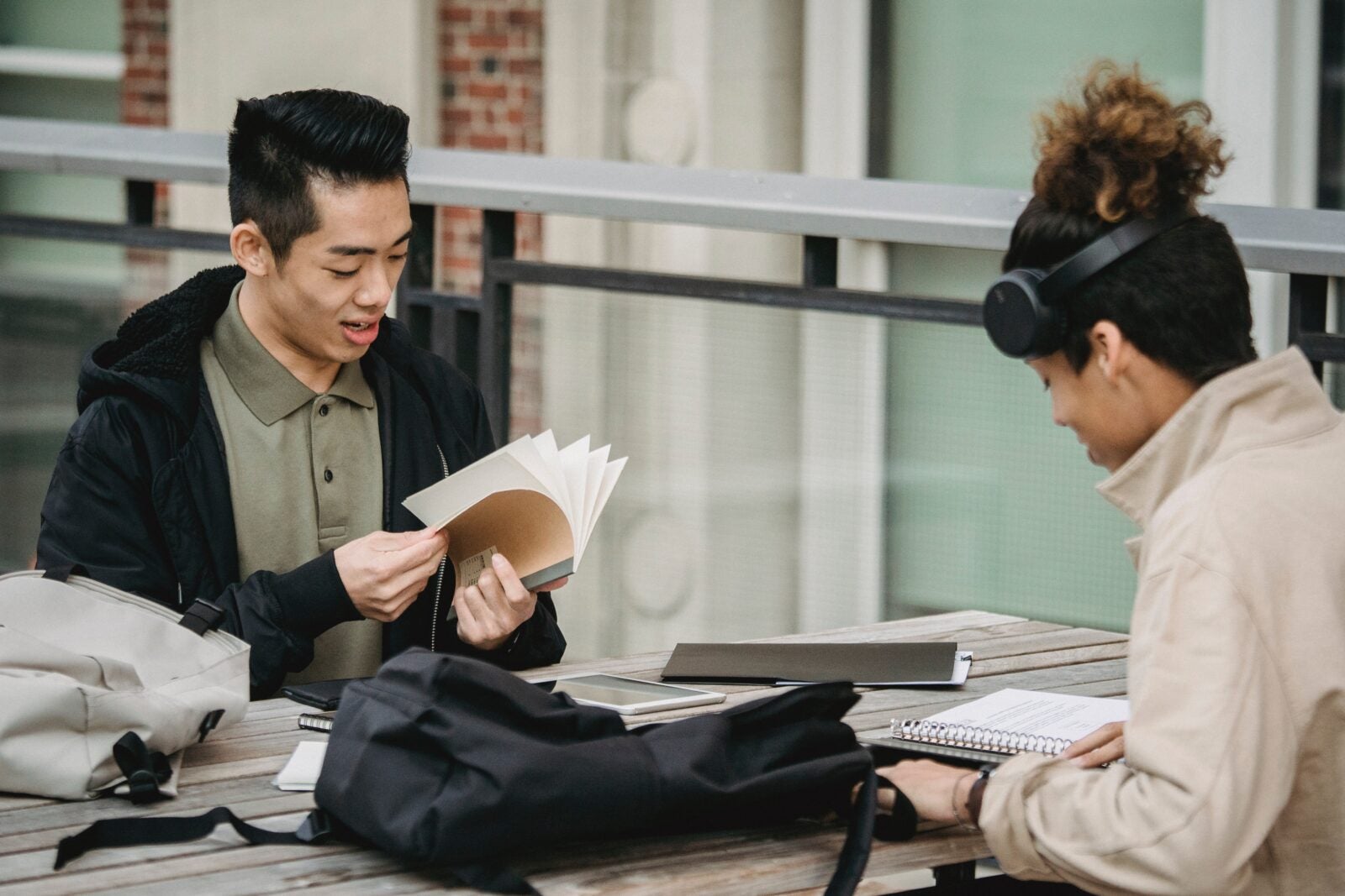 A young Asian man flips through a book while sitting at a table with a woman with curly hair and dark skin. 