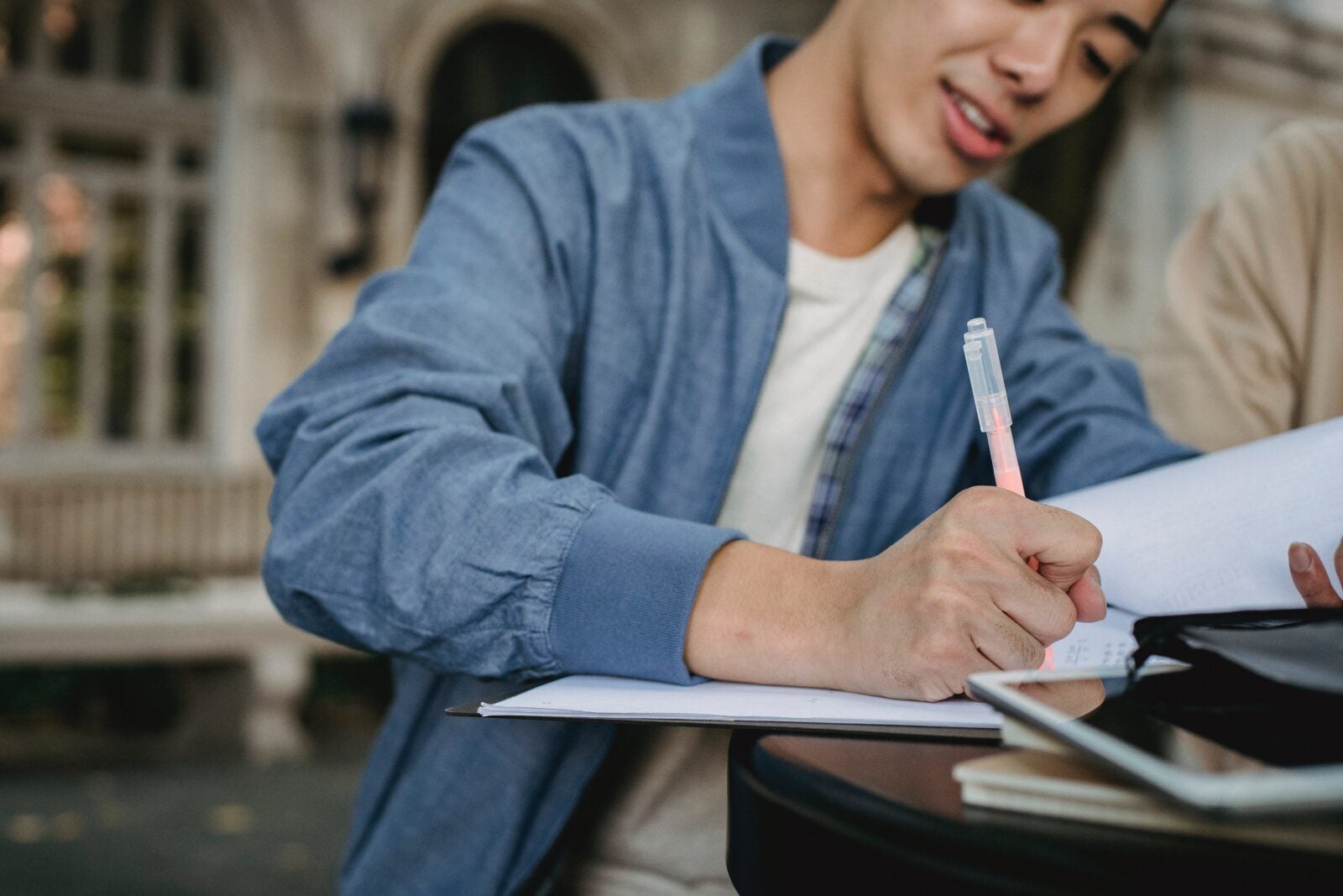 A young Asian man wearing a blue bomber jacket writing in a notepad using a colored pen.