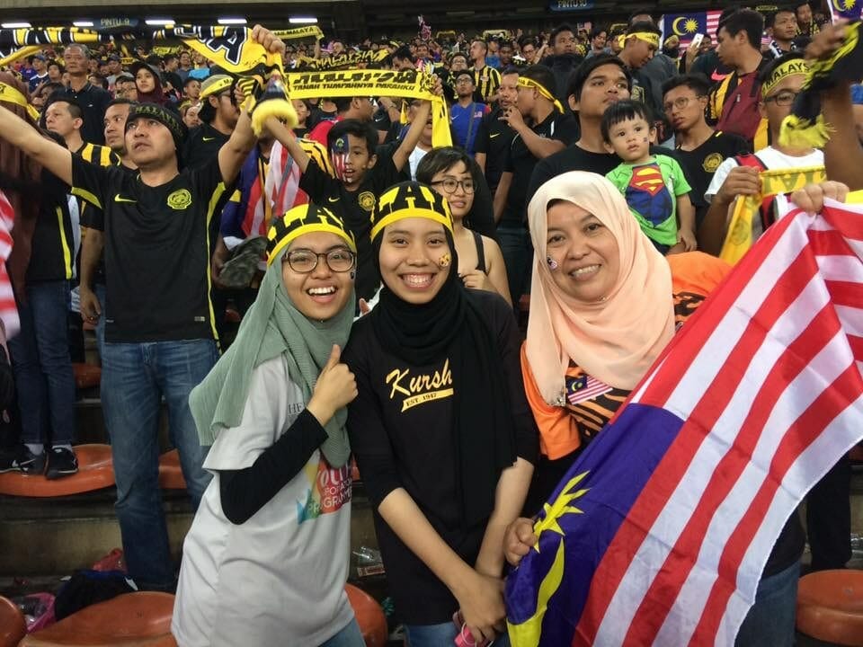 People cheering and waving the Malaysian flag during a sporting event.