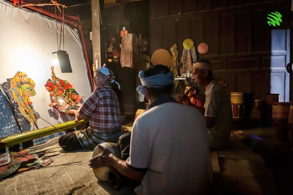 A Practitioner Of Wayang Kulit Sits Down In His House Surrounded By His Craft.