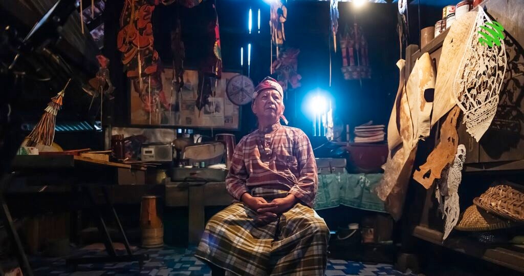 A Practitioner Of Wayang Kulit Sits Down In His House Surrounded By His Craft.