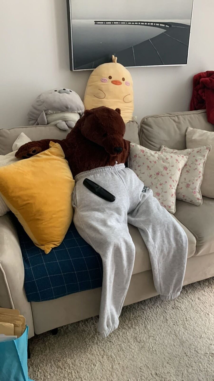 Ikea's Djungelskog bear wearing a grey sweatpants, sitting on the sofa with pillows on either side of it. A television remote control sits on its lap.