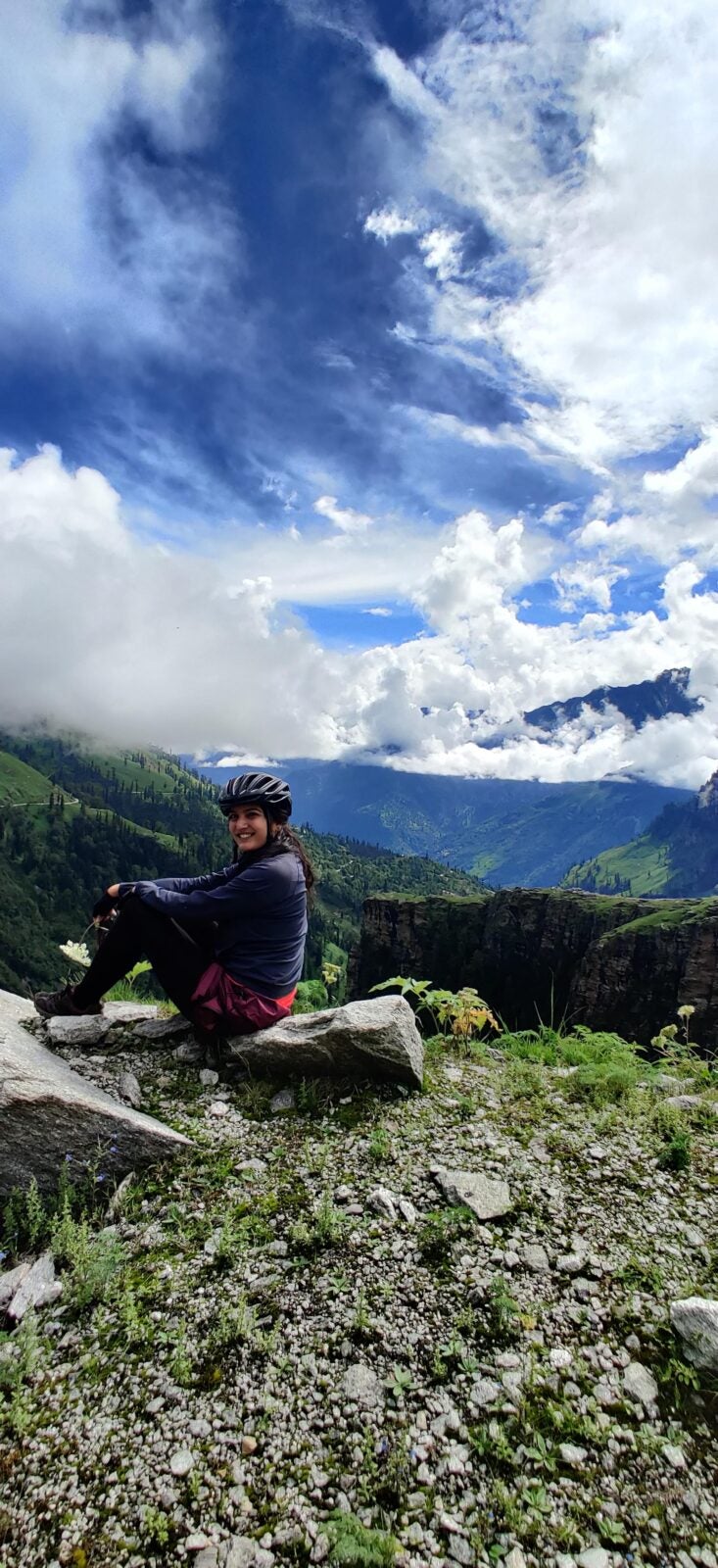 Pashmina wearing her cycling helmet and sitting on a rock while smiling. Behind her are mountains and clouds.