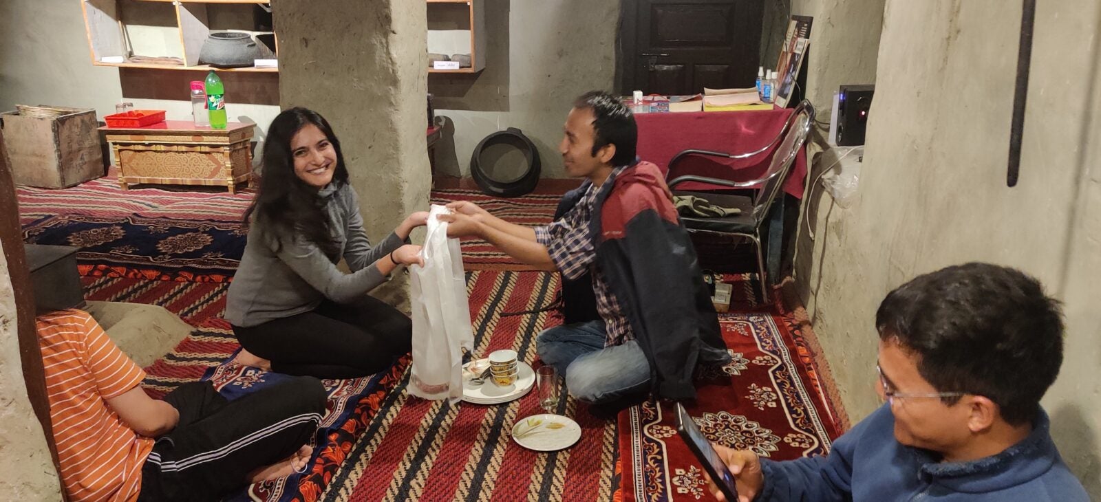 Pashmina accepting a goody bag from a man while they sit on the floor.