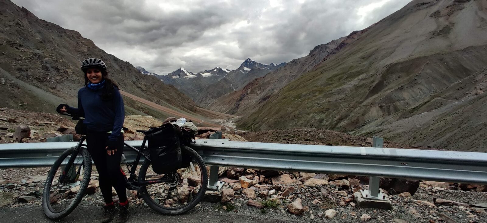 Pashmina standing next to her bicycle at the side of a road. Beyond her are mountains and a very cloudy sky.