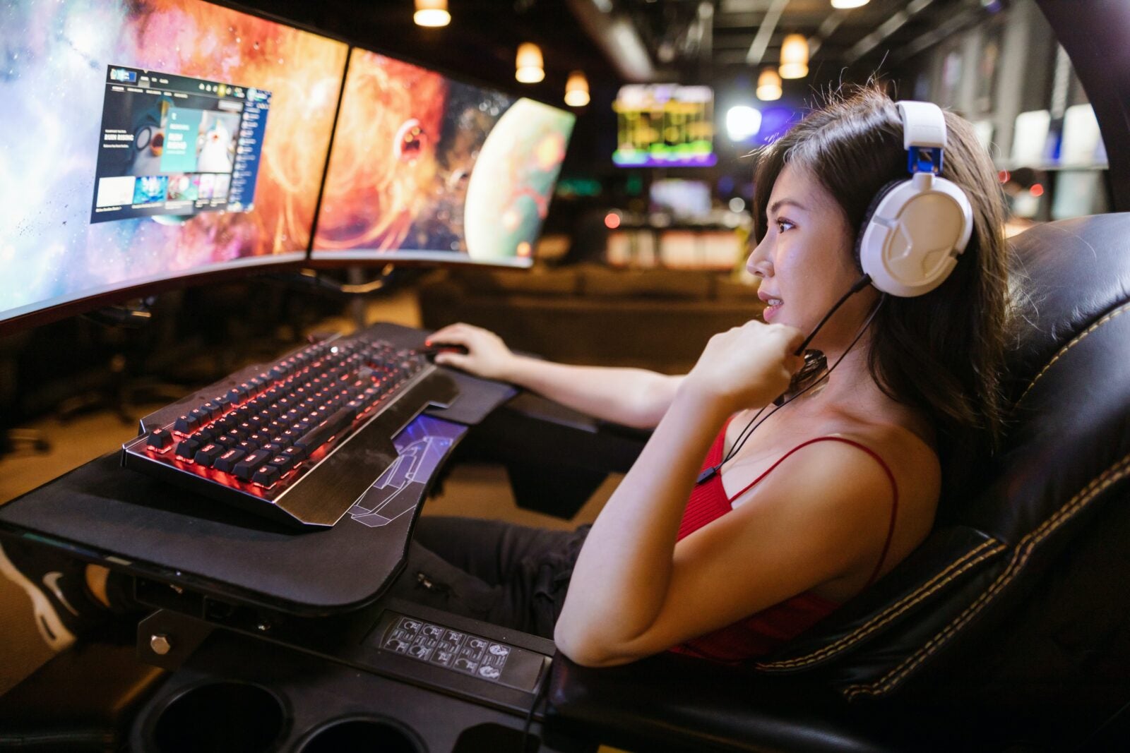 An Asian woman wearing a red top and white headphones using a gaming computer.