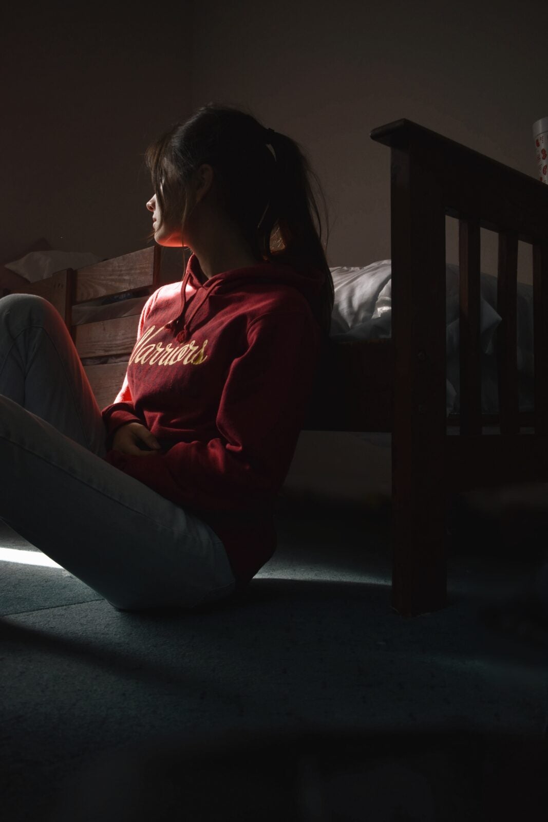 A woman wearing a red long sleeve shirt and white pants, sitting on the floor by the bed while looking out towards a light source.
