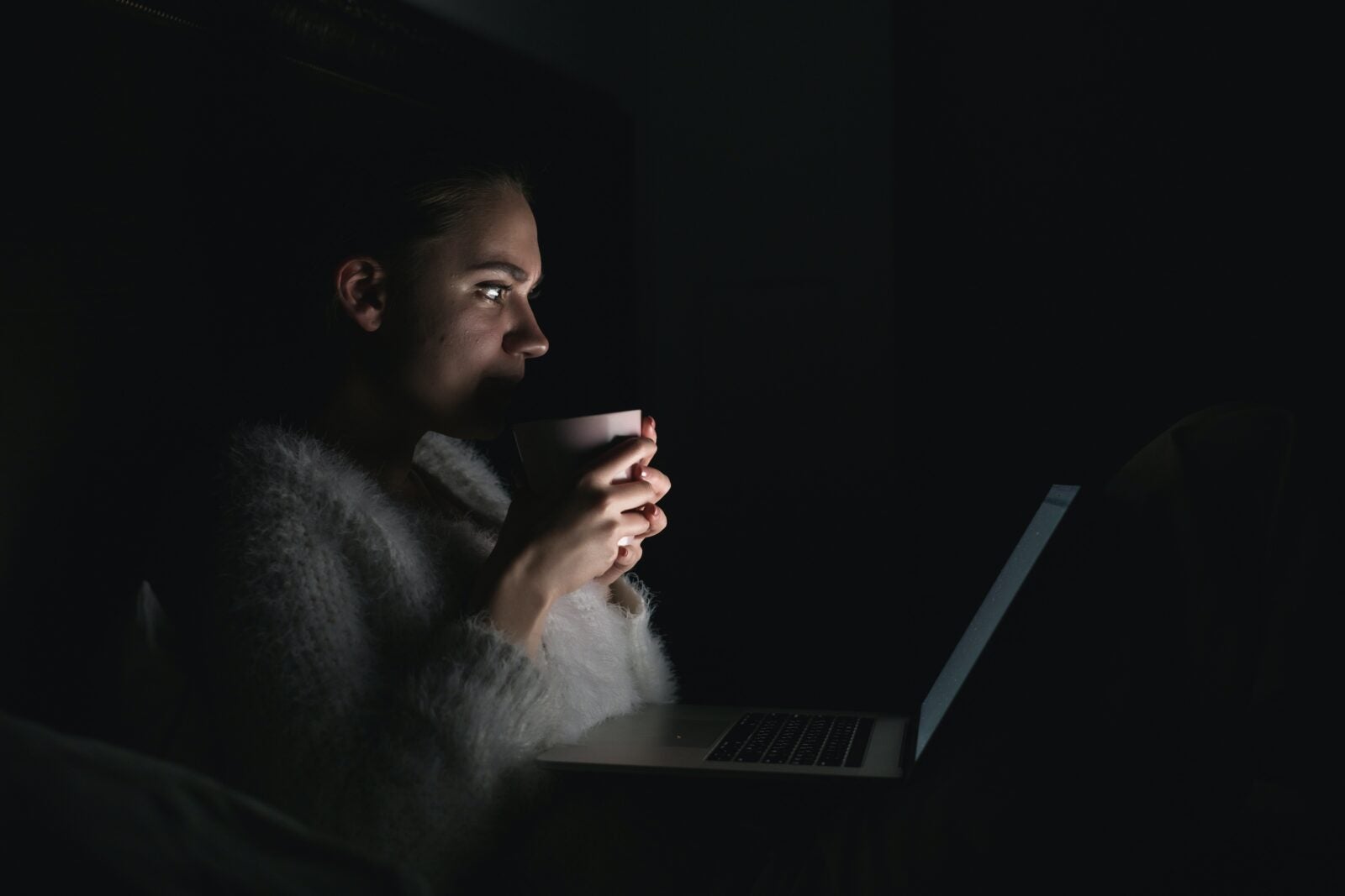 A woman sitting in the dark and drinking a cup of glass in front of her laptop.