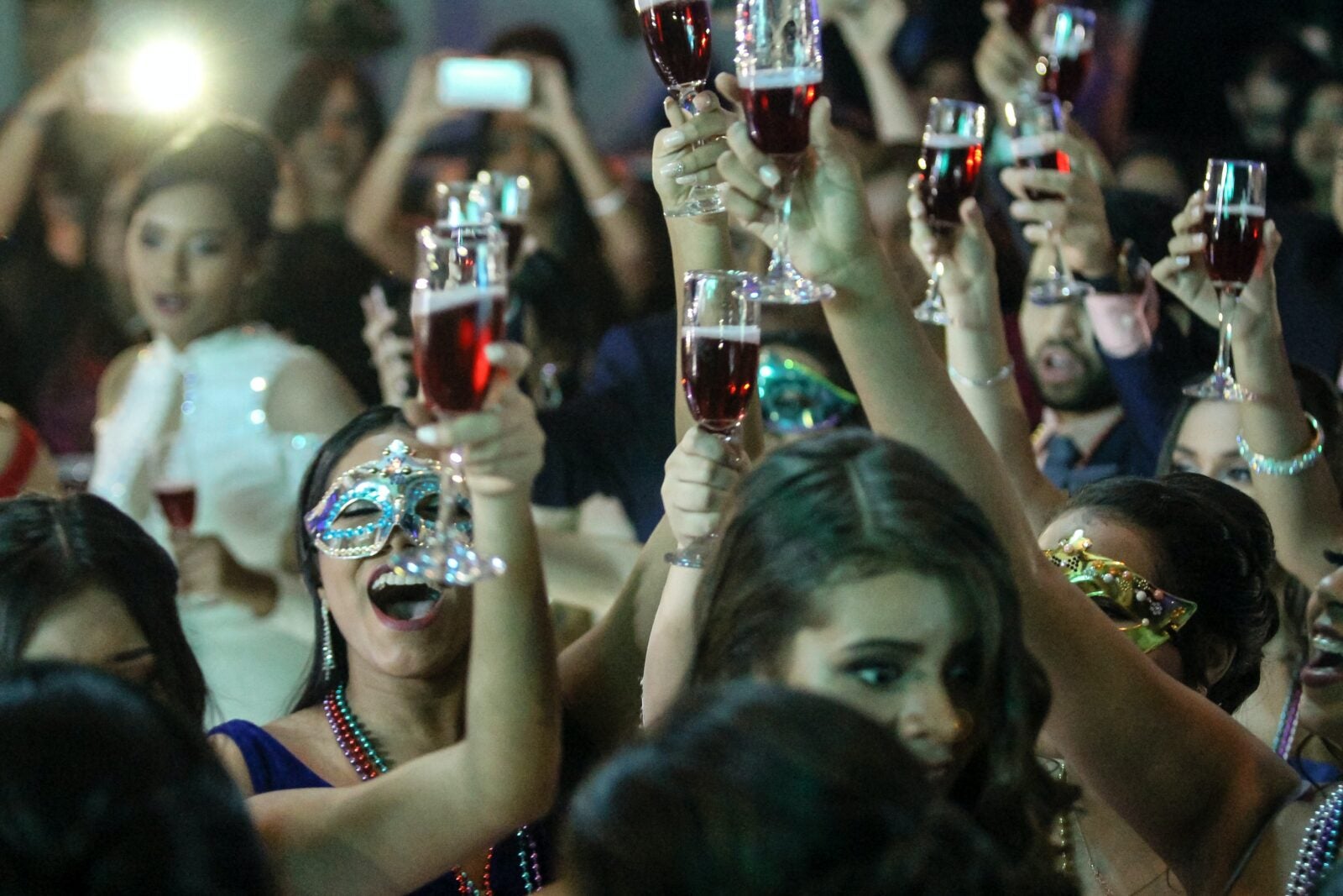 A party where some girls are wearing decorative masks and holding a glass of red liquid in the air.