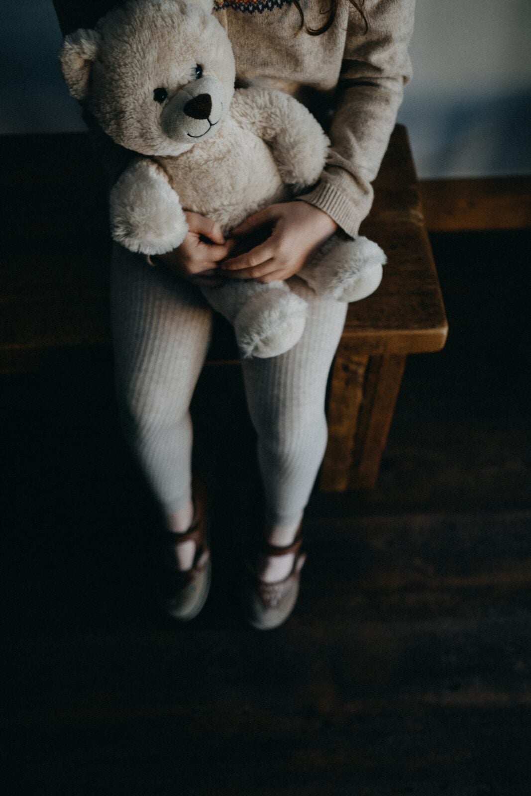 A child wearing ribbed leggings sitting on a wooden bench and holding onto a white teddy bear 