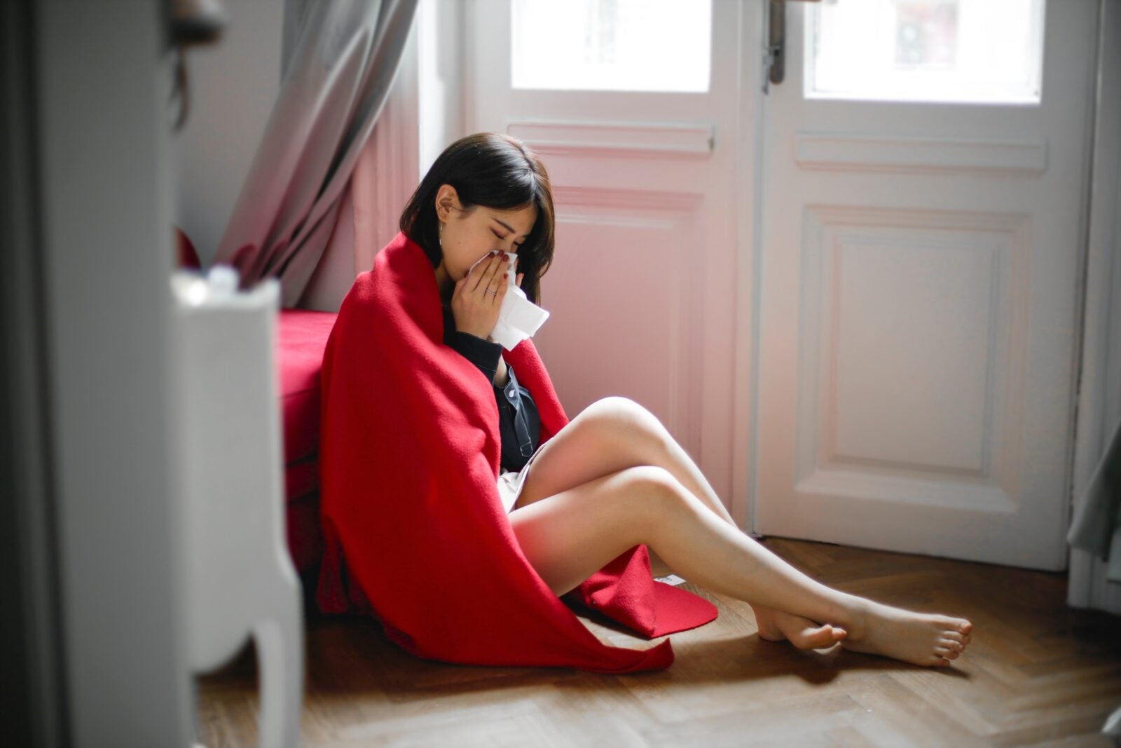A young Asian woman sitting on the floor with a red blanket over her shoulders and wiping her nose.