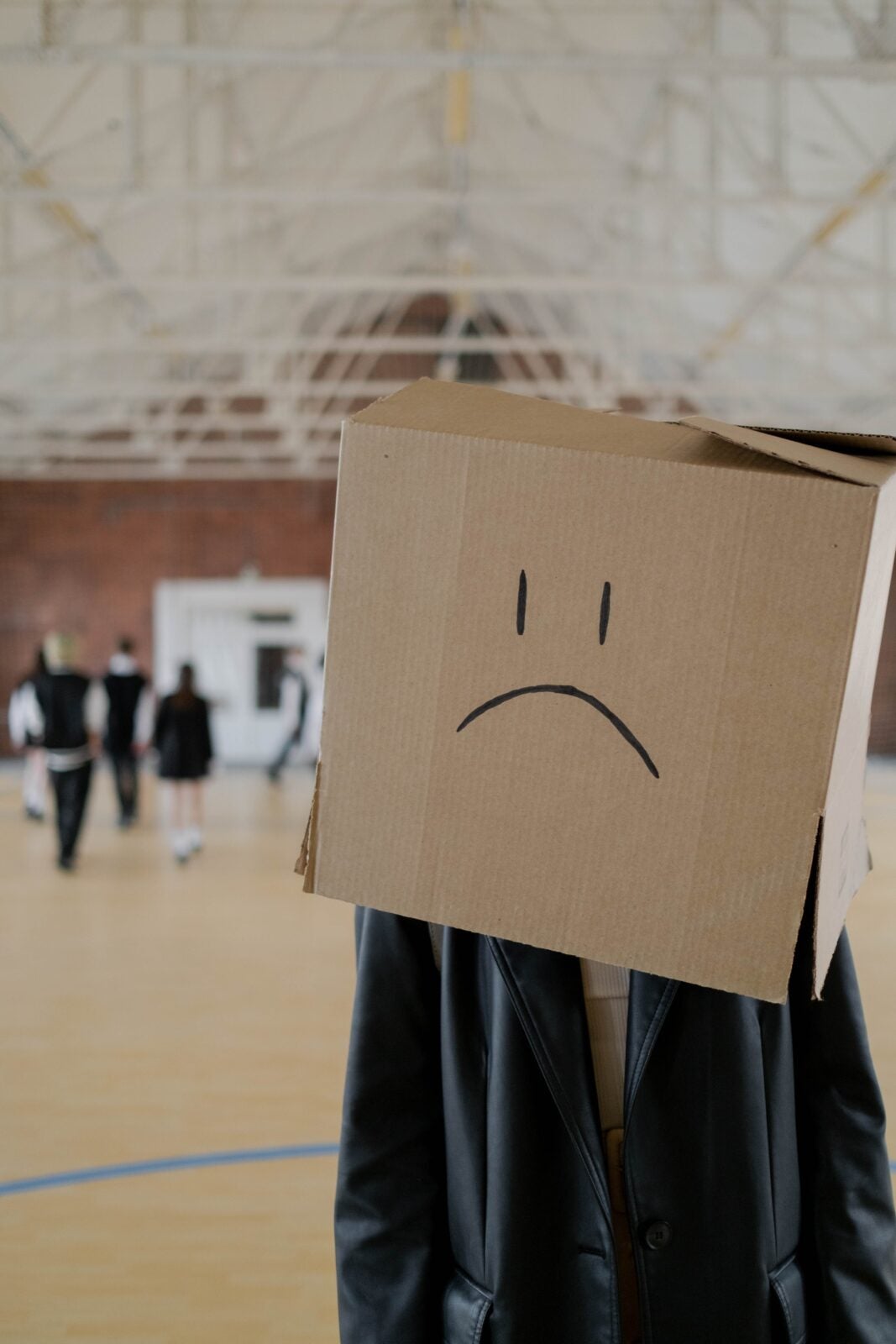 A man with a box over his head standing in an indoor school gym. The box has a sad face drawn on it.