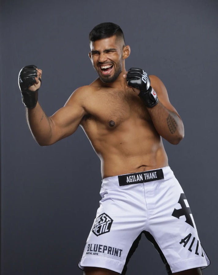 Agilan Thani, a boxer posing shirtless with gloves on and smiling.