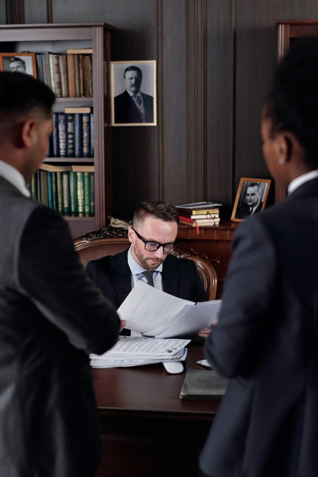 A man sitting behind a desk looking at several pieces of paper while two men facing him in front of the desk. They are wearing suits.