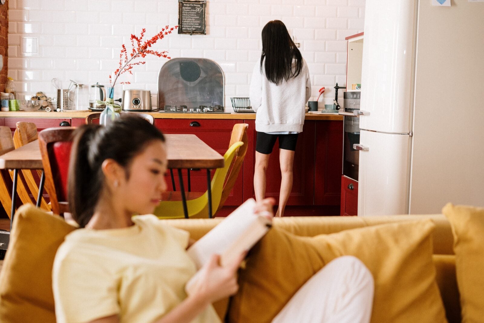 An Asian woman sitting down and reading a book while another woman is in the kitchen.