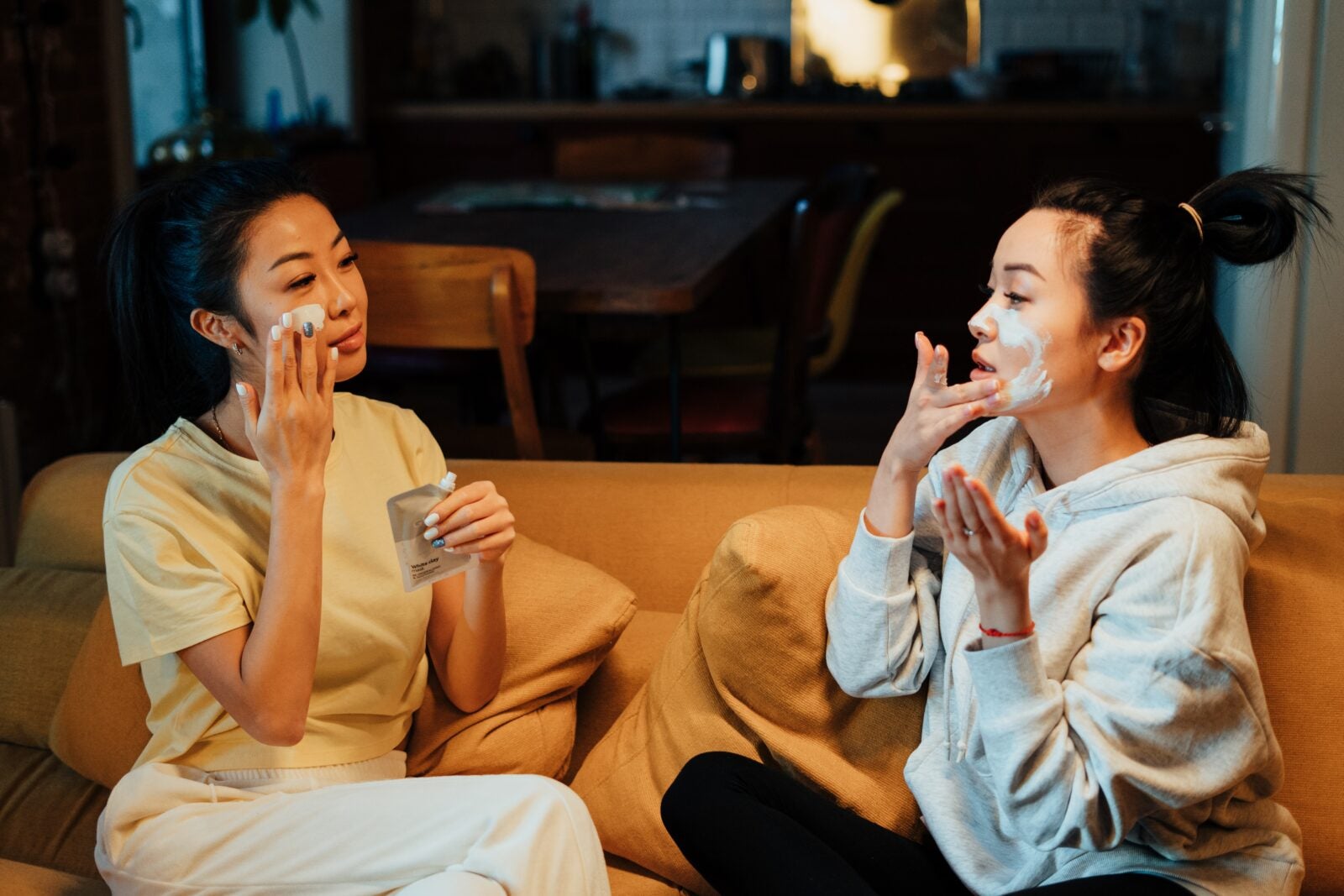 Two Asian women sitting on a sofa in their living room at night and putting on a facial mask.