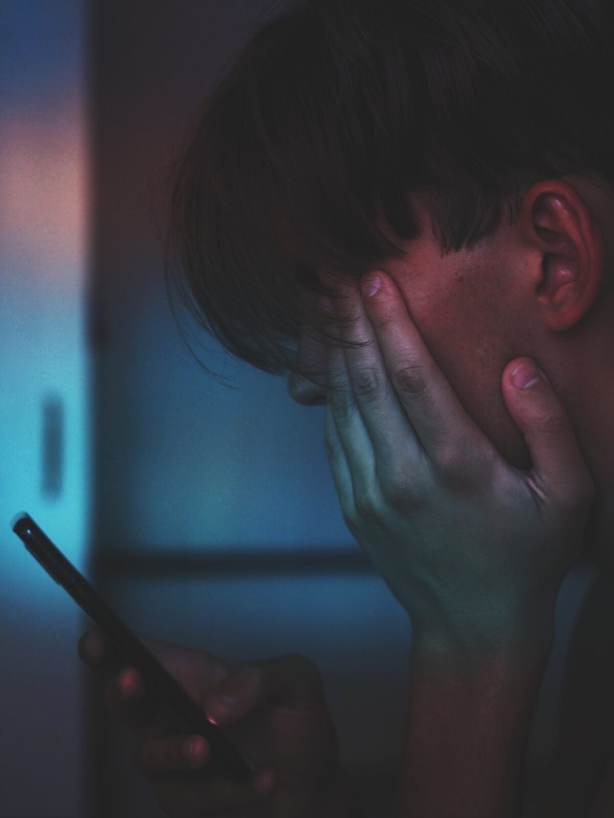 A man with one hand covering a side of his face while he uses his phone in a dark room.