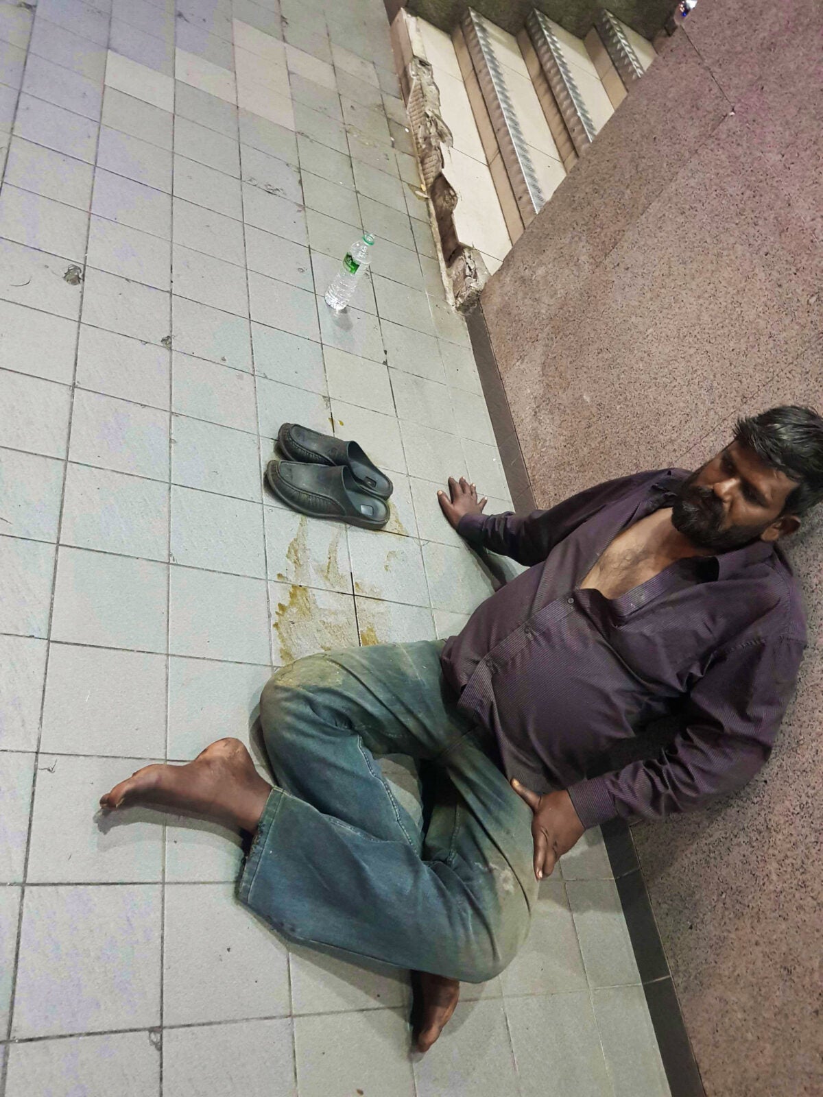 A Homeless Malaysian Man sitting on the floor which is stained with brown liquid and leaning on the wall .