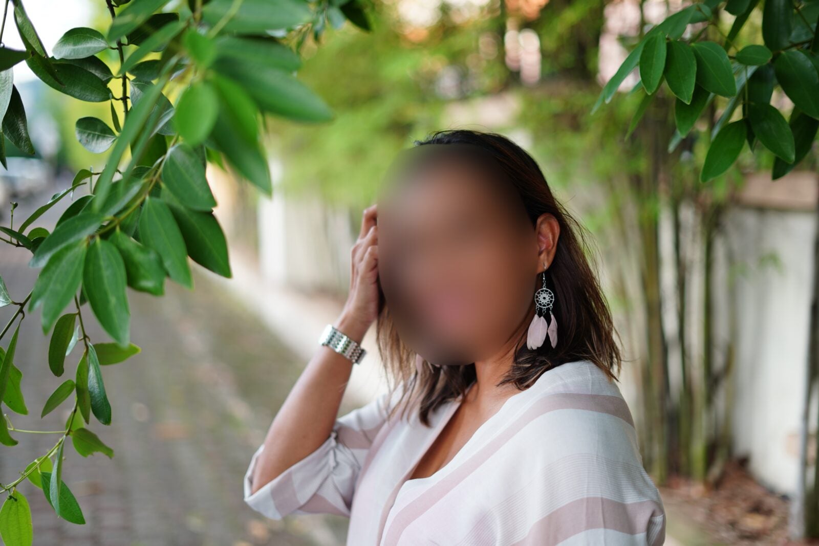 Woman in white blouse and dangling earrings standing under a tree on the sidewalk of a street.