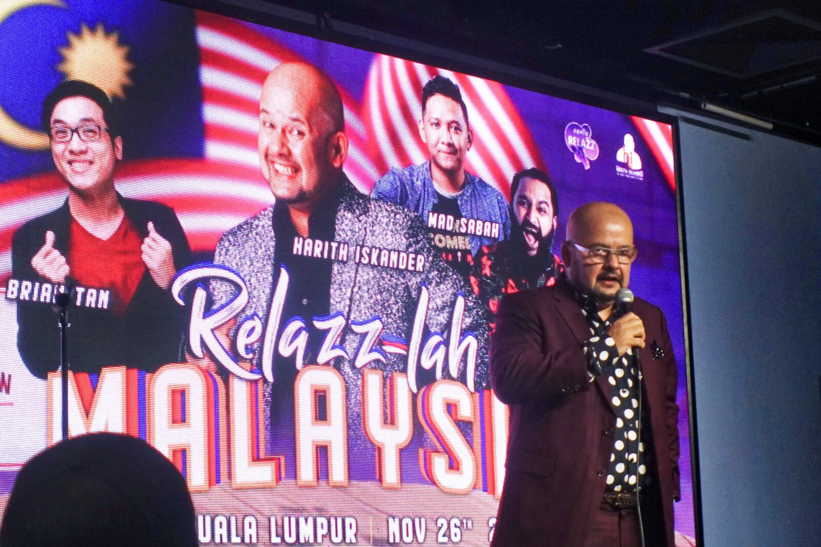 A Bald, Well-Dressed Man Stands On Stage Holding A Microphone. Behind Him Is A Screen That Showcases An Upcoming Event On The 26Th Of November Called &Quot;Relazz-Lah Malaysia&Quot; Which Features Four Comedians: Brian Tan, Harith Iskandar, Mad Sabah And Prakash Daniel.