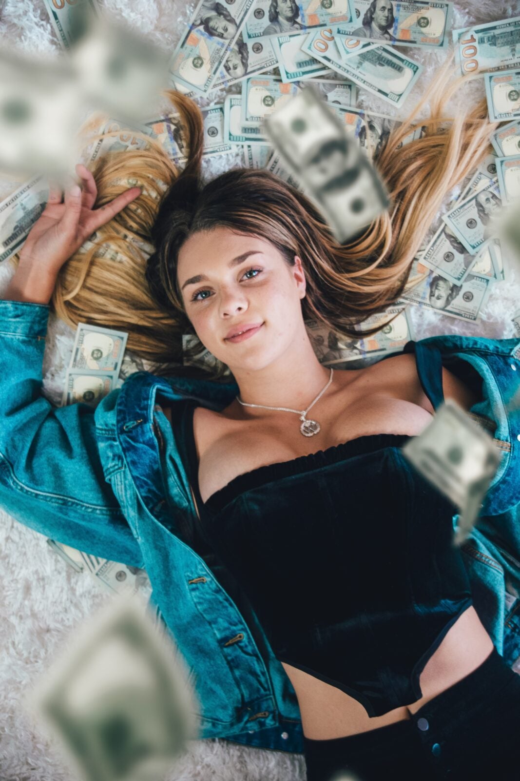 Young woman wearing a corset and jean jacket laying on top of a bed of money.