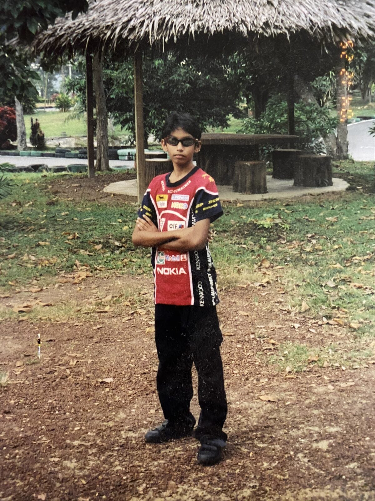A Malaysian Indian boy wearing sunglasses stands in a clearing of a garden with his arms crossed together.