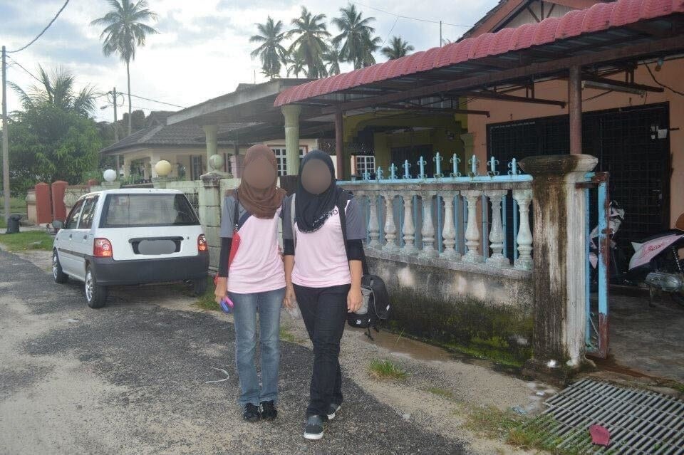 Two Malay women wearing the hijab standing close together in front of a home with moldy concrete fences and gate.