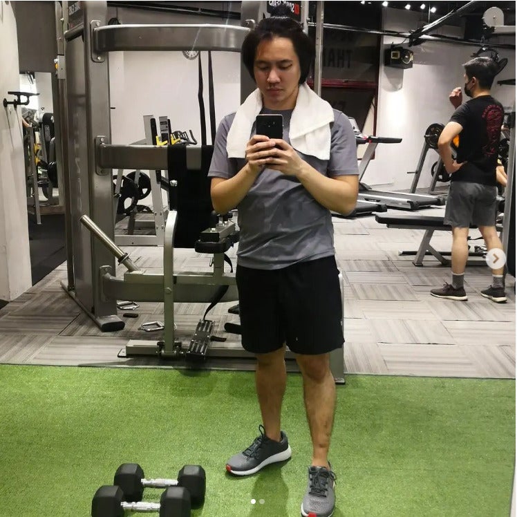 A man wearing shorts and a shirt with a towel resting around his neck taking a selfie with his phone at the gym surrounded by gym equipment and other gym gooers.