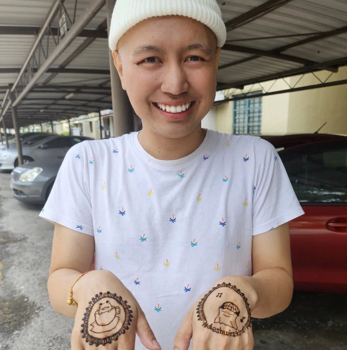 A Malay man wearing a beige beanie smiling and showing the henna designs that are drawn on his hands.