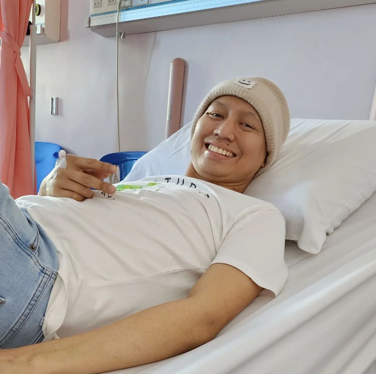 A Malay man wearing a beige beanie lying on a bed at a hospital with a butterfly needle poked into his hands for an IV drip.