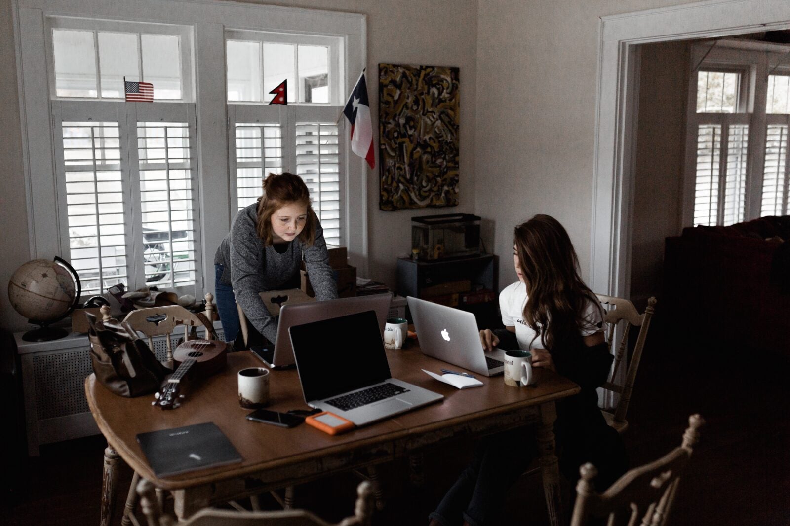 Two girls sitting in the dining room of their home and doing work on their laptops.