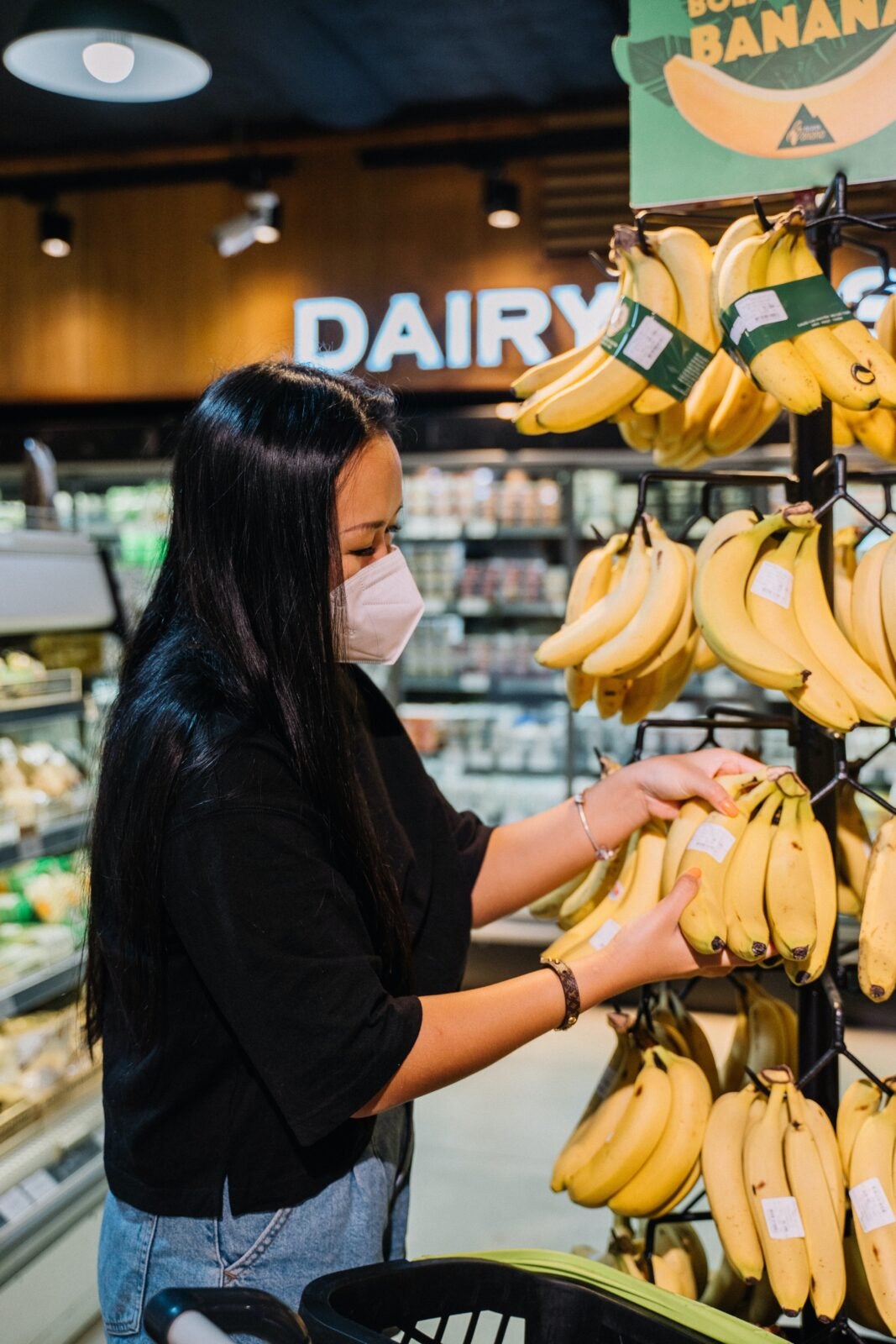 A masked Asian woman picking bananas at a grocery store.