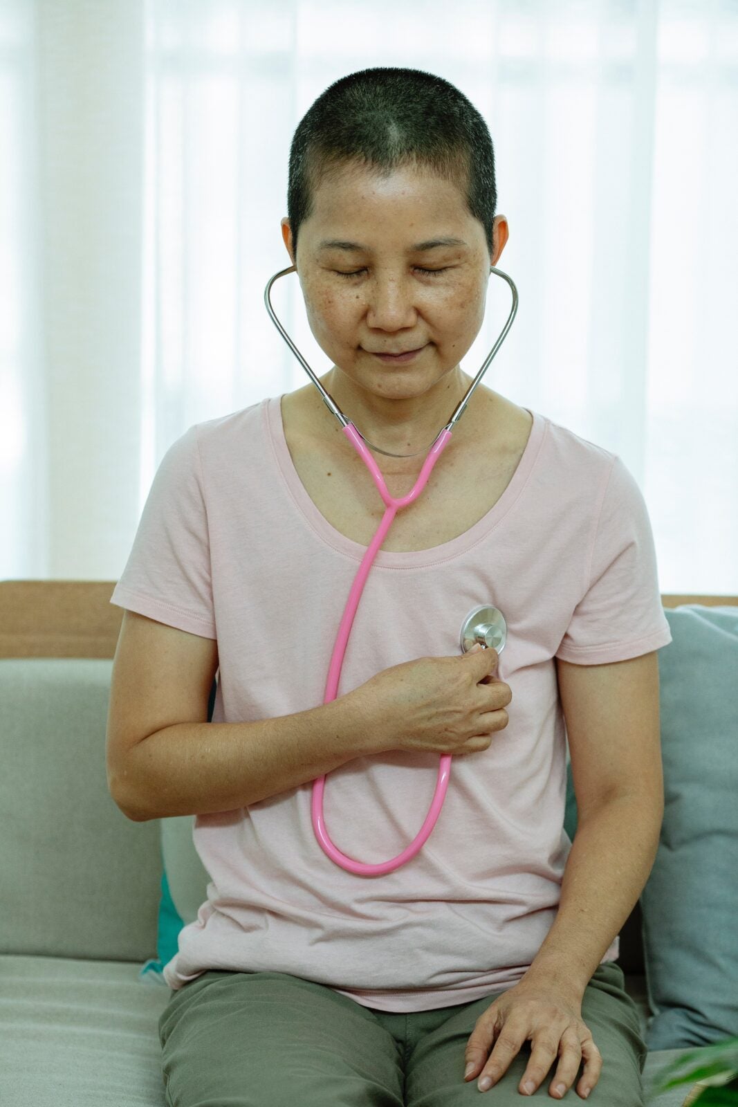 A bald Asian woman holding a stethoscope to her own chest.