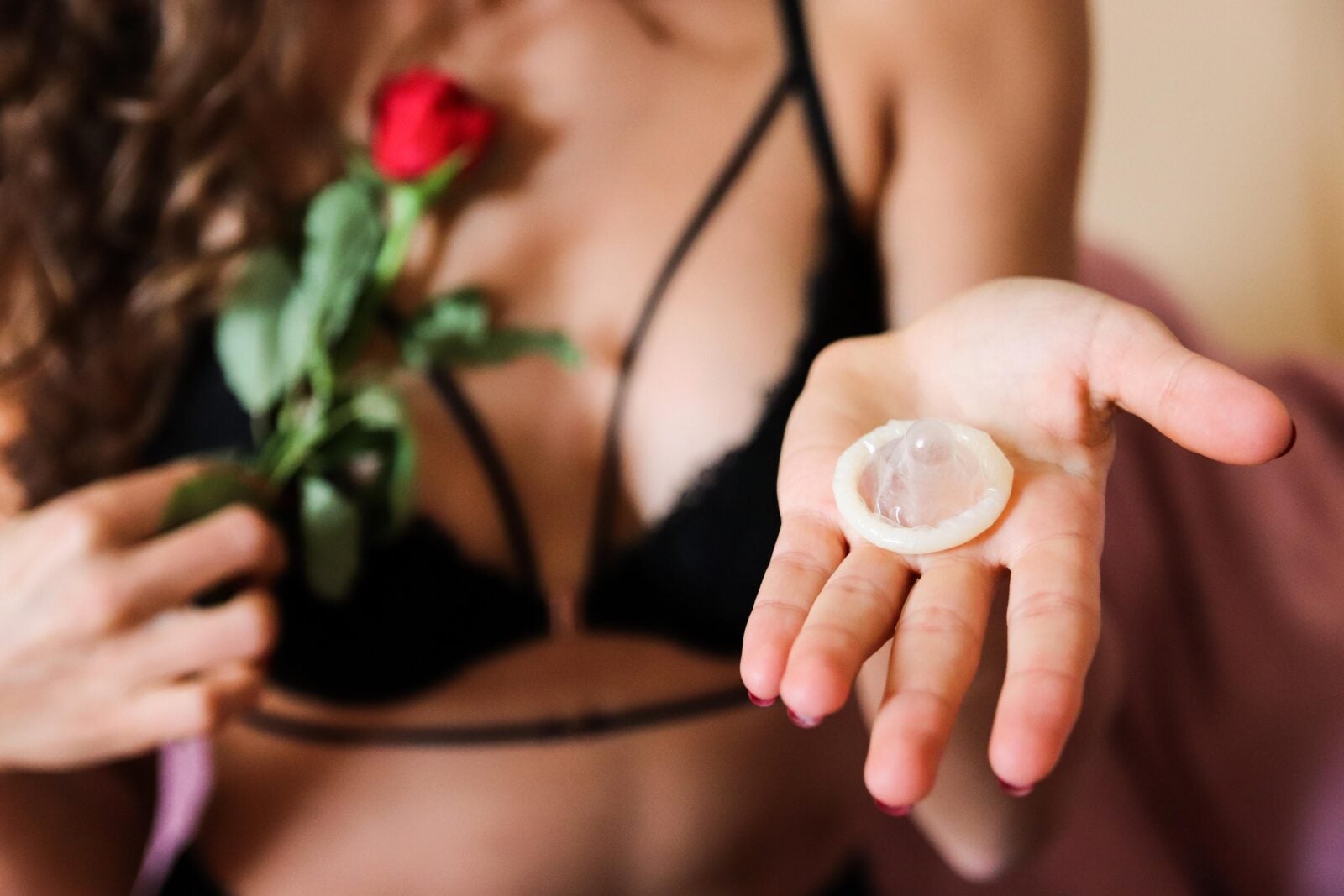 Woman wearing black bra and holding a red rose in one hand and a condom in the other hand.
