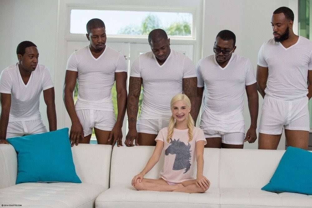 A blonde white woman sitting on a white sofa while five black men stand behind the sofa while looking over her. The men are wearing white boxer briefs and white tight shirts while the woman is wearing a shirt with a unicorn graphic on it.