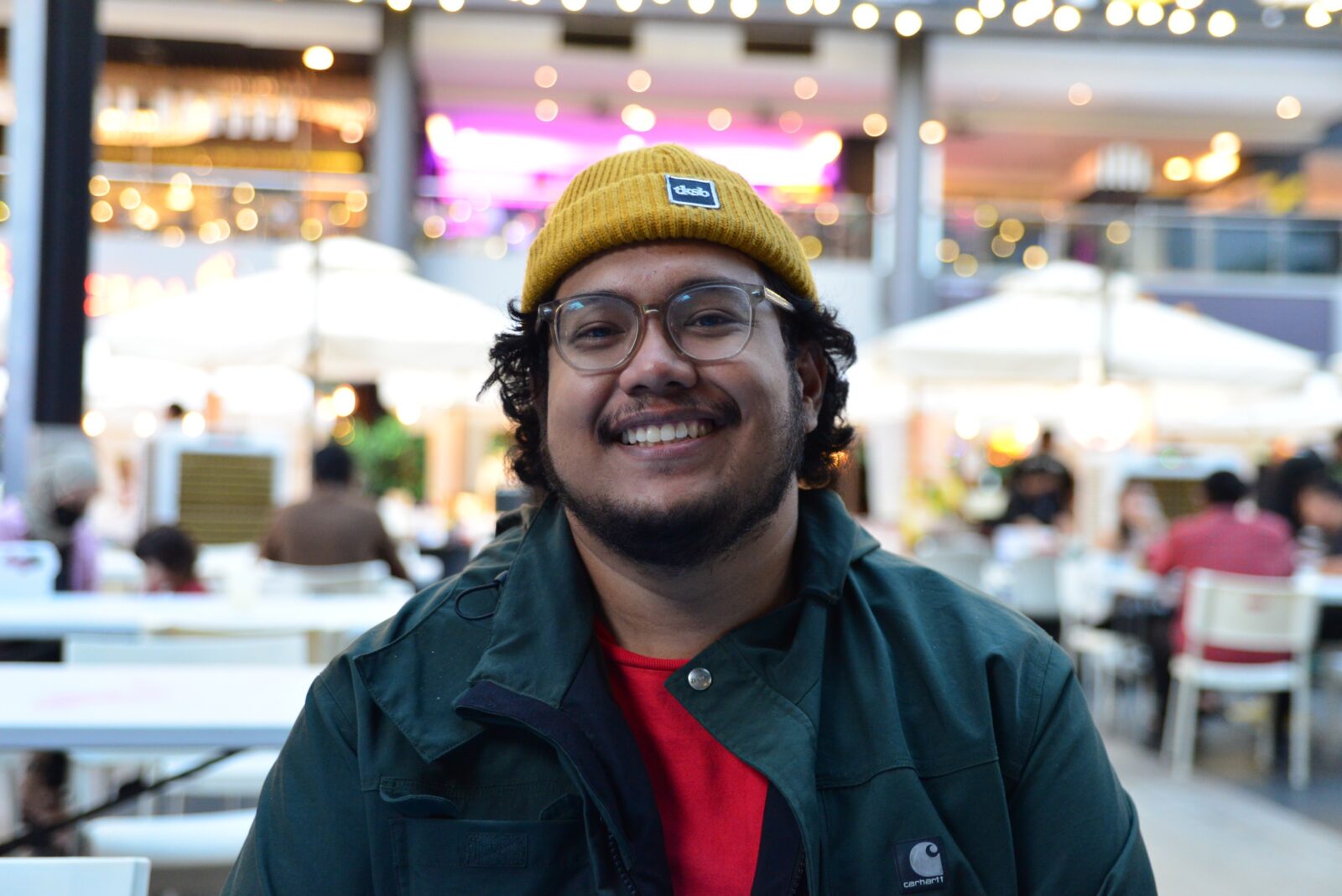 A man wearing a yellow beanie, green jacket and red shirt with glasses on while smiling. 
