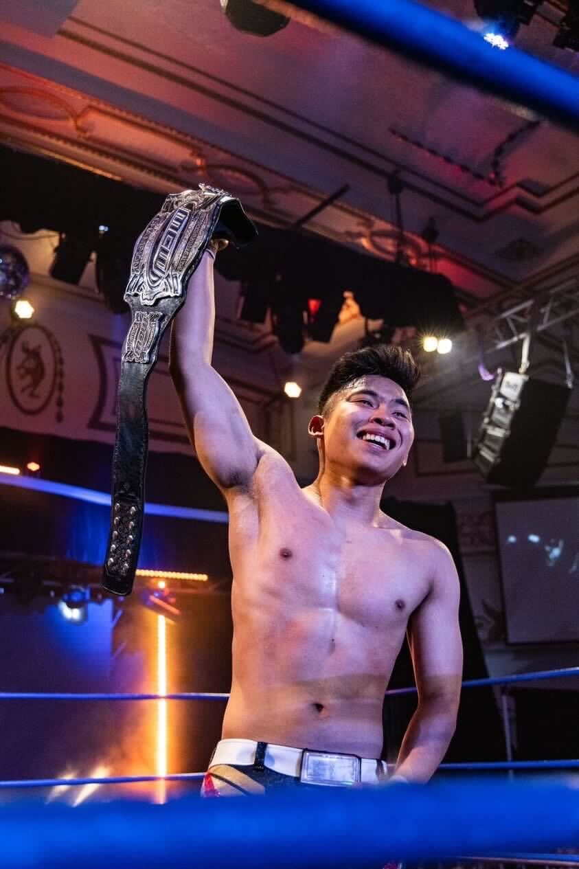 Young Malay man holds his championship wrestling belt high above his head and smiles with glory.