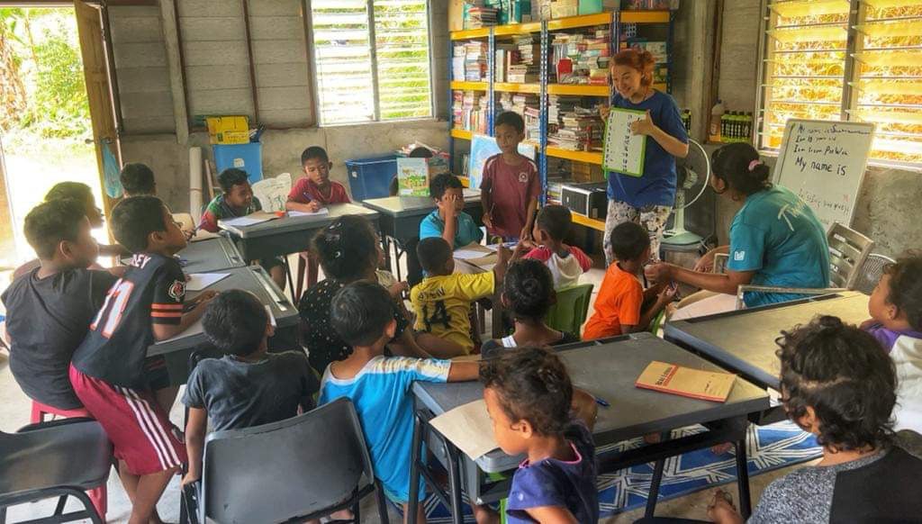 A classroom full of Semai children of different ages and a woman who is their teacher holding a visual aid for teaching.
