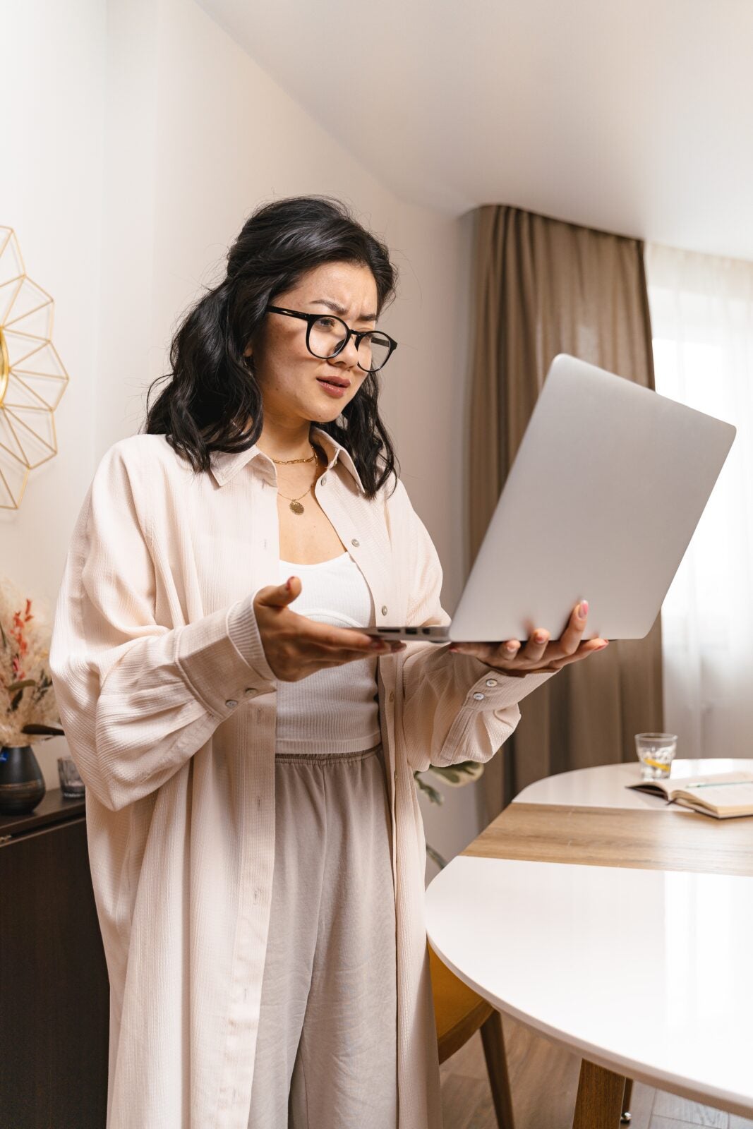 Asian woman wearing glasses, a long shirt and loungewear looking at her laptop with furrowed eyebrows.