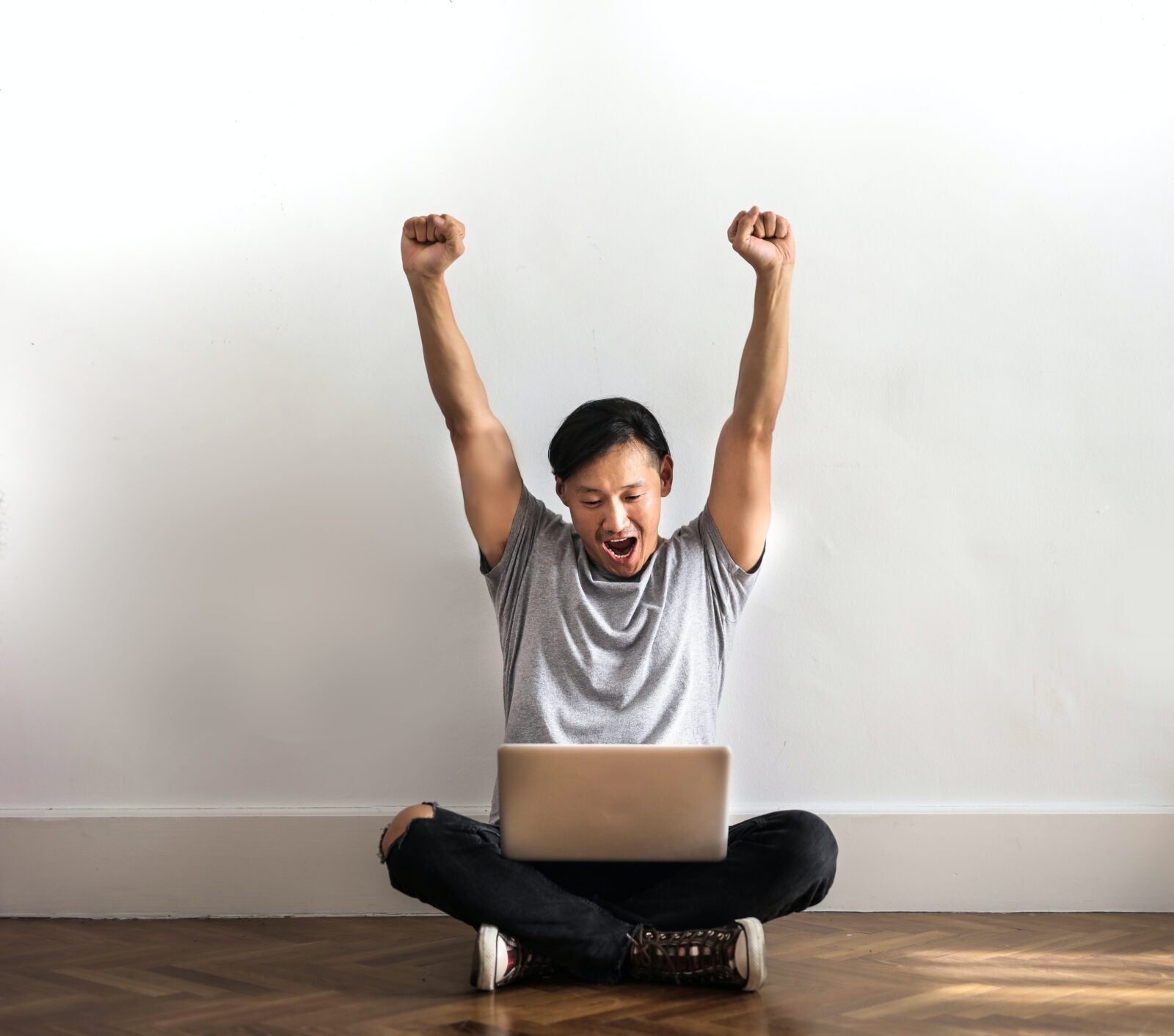 Asian man sitting on the floor using a laptop and holding his hands above his head in victory.
