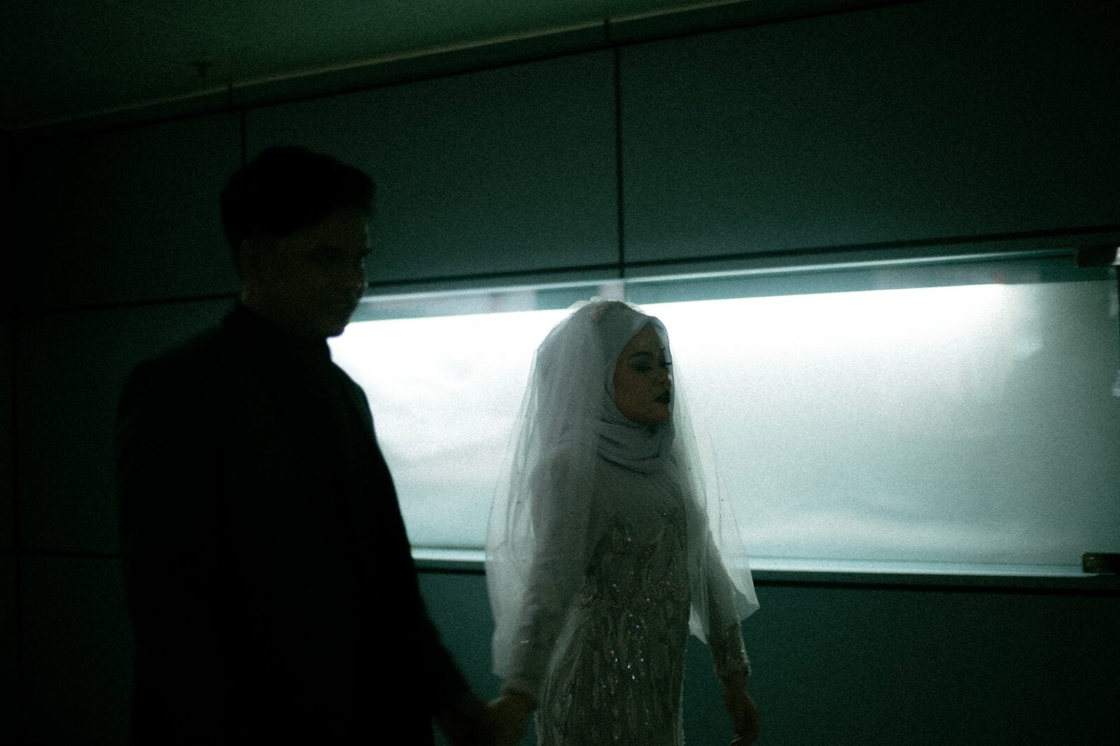 A Malay woman wearing a white wedding dress, white hijab and a white veil walks alongside her husband who is casted in a shadow along a white lit hallway.