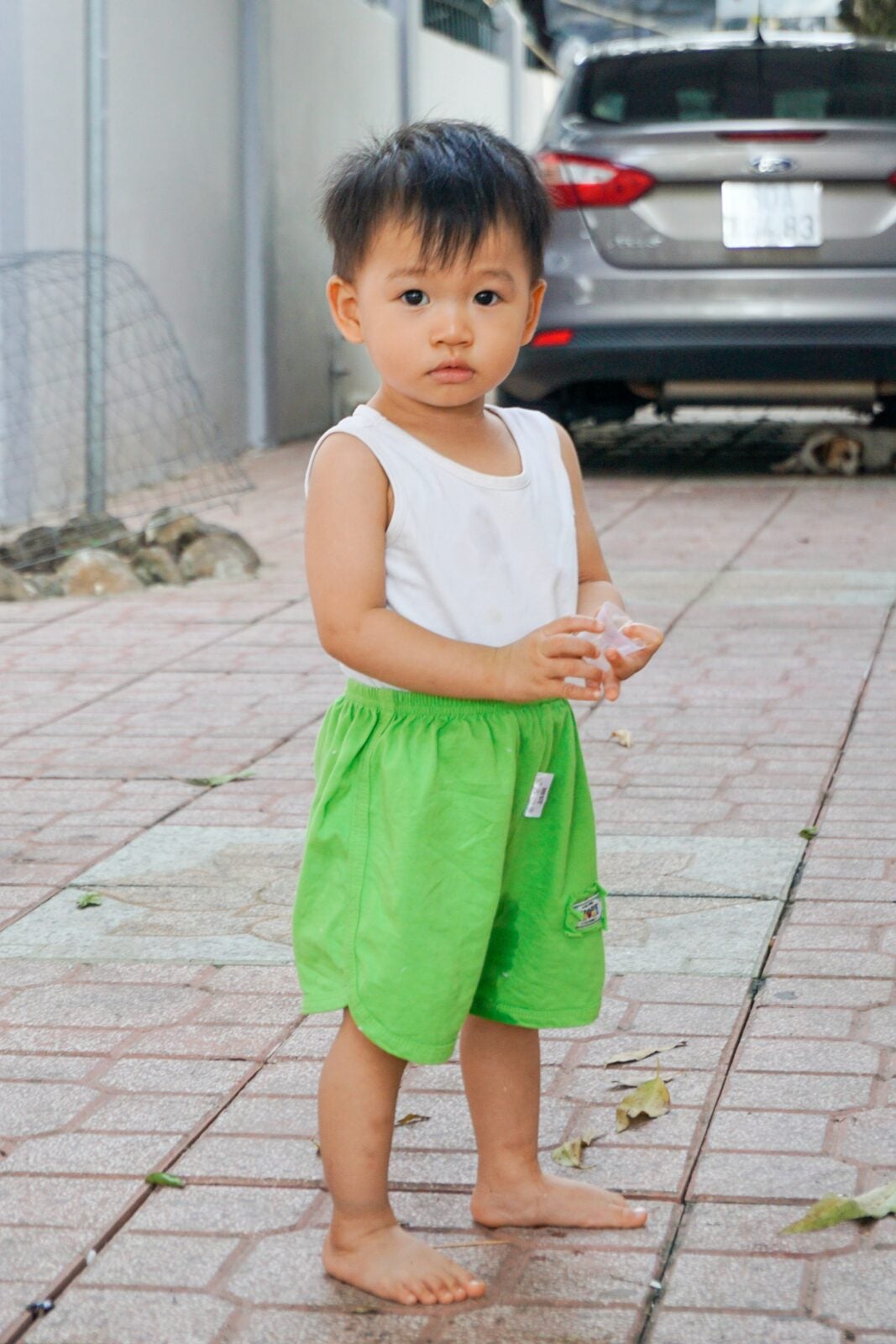 A young Asian boy in green shorts and white tank shirt standing in a driveway holding a plastic cup.