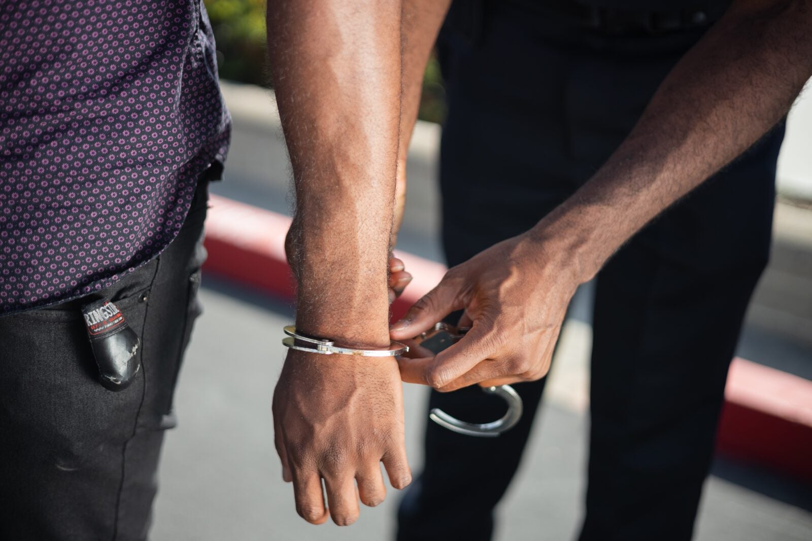 A dark skinned man's hands being hand cuffed by another dark skinned man.