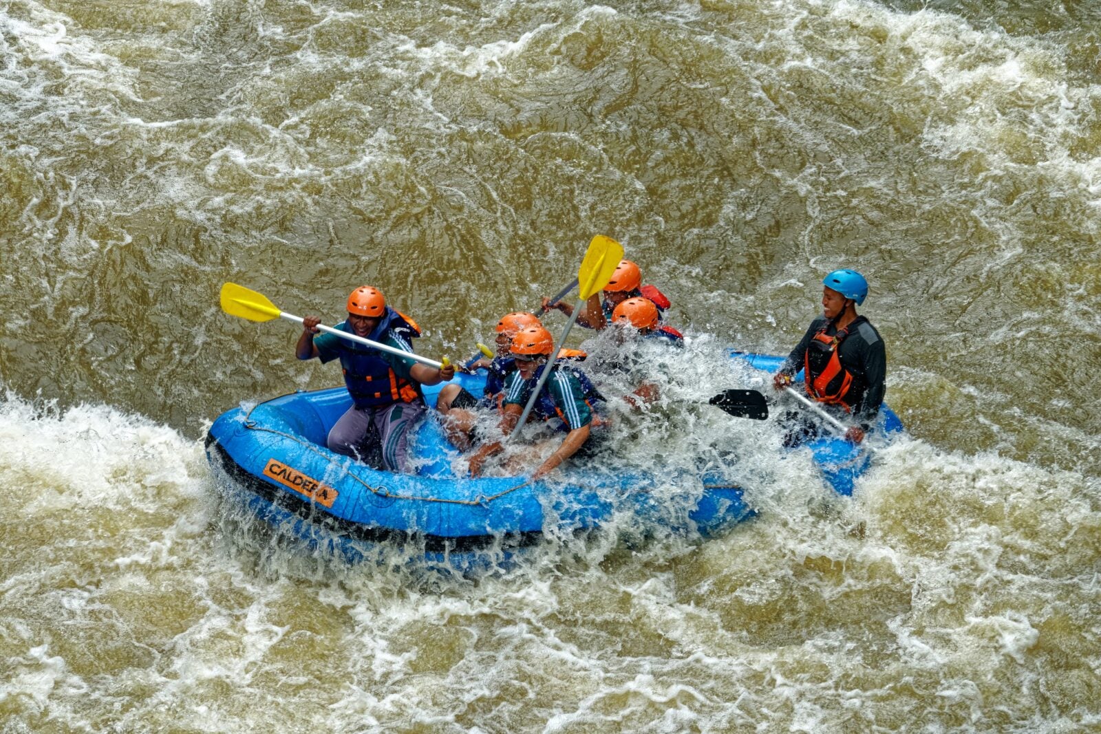 Four men wearing bright colored helmets in a blue raft paddling in rough waters