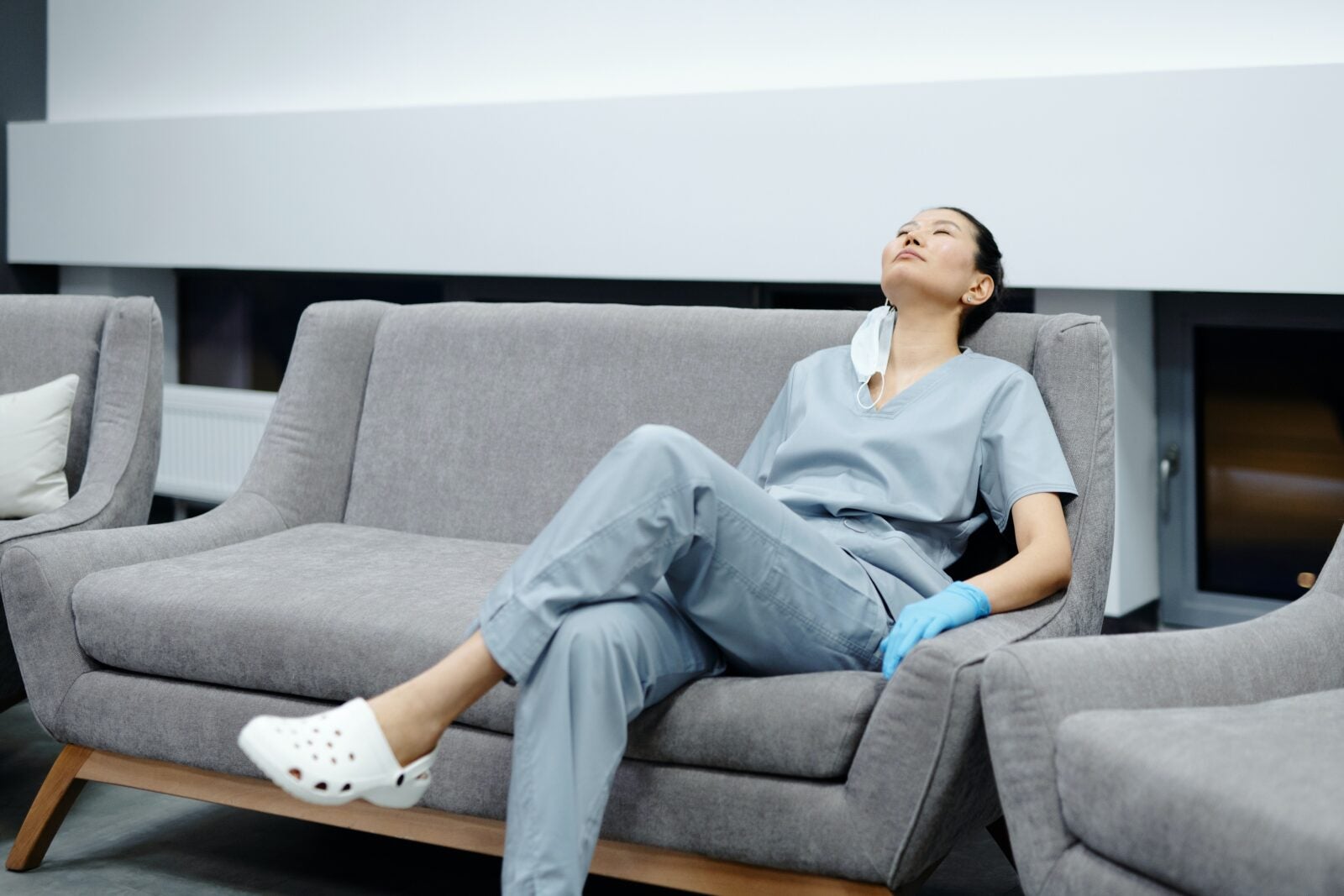 A young Asian woman wearing hospital scrubs, white crocs and blue gloves sit on a 2 seater sofa while resting her head and crossing her legs.