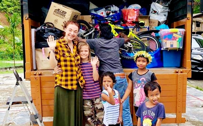 A ginger-haired woman waves together with her son and three young indigenous children. They stand in front of a truck that is being loaded by a man.