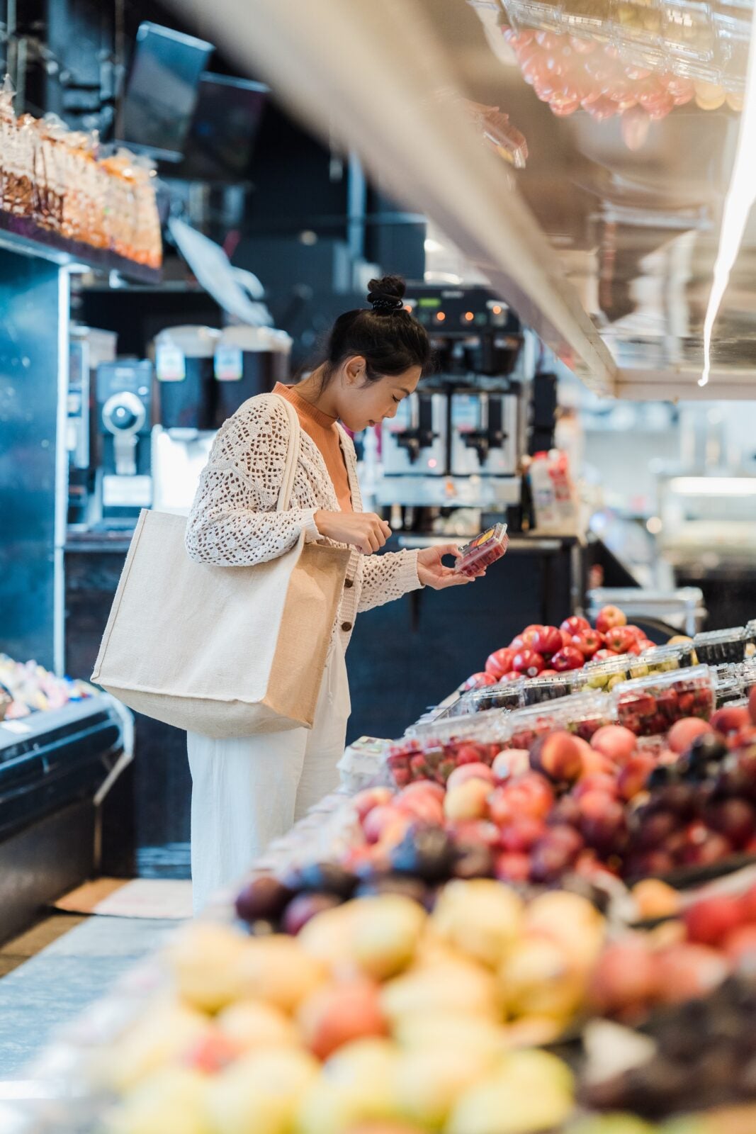 An asian woman holding a square white bag while looking at the fruits in a grocery store.