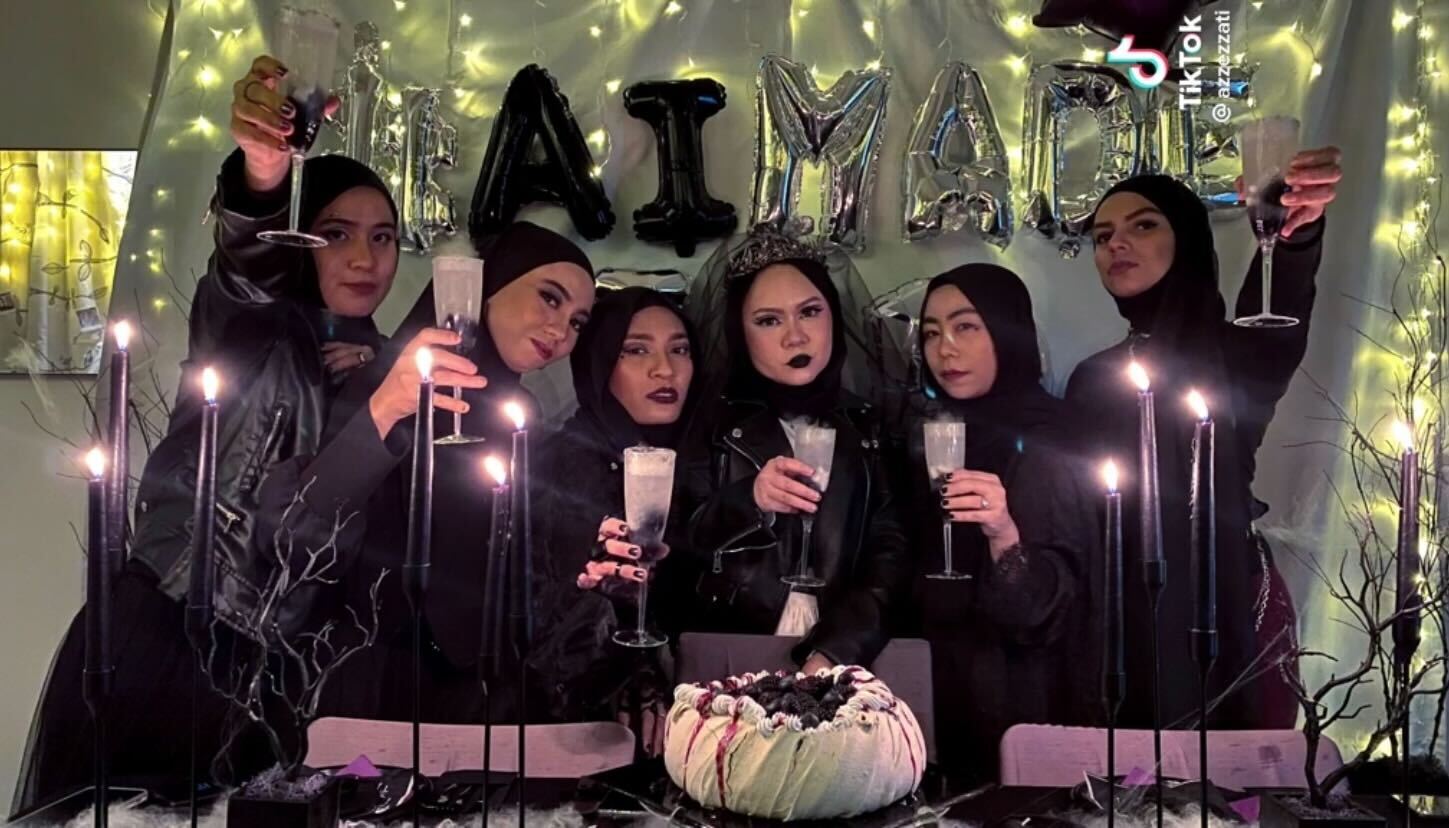 Six gothic women in hijab posing in front of a pavlova cake with black frosting while hoding a fancy drink and alphabet balloons behind them that spells: # AI Made It.