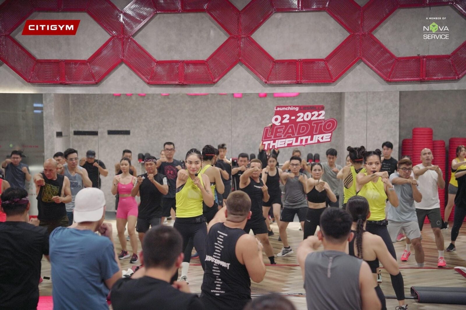 A group of people doing fitness exercises with the help of an instructor at Les Mills in 2022 with the slogan "Lead to the future" printed on the wall.